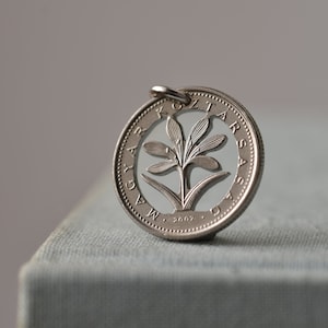 Pode incluir: Pingente de prata feito a partir de uma moeda, com um design floral recortado. A moeda tem texto ao redor da borda e o ano de 2002. O pingente é preso a um pequeno laço de prata para pendurar.