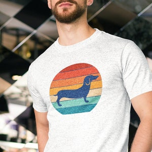 May include: A light grey t-shirt with a retro-style dachshund silhouette in a circular design. The circle features a gradient of colours, including red, orange, yellow, blue, and teal. The t-shirt is made of a soft material.