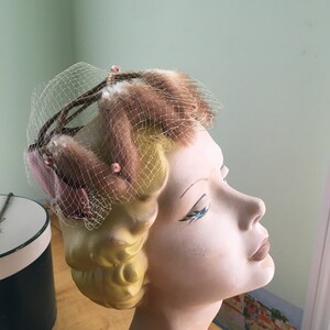 Puede incluir: Un sombrero vintage con una red marrón y rosa y plumas marrones. El sombrero está en una cabeza de maniquí con cabello rubio.
