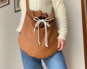 May include: A brown leather tote bag with a drawstring closure and white cotton cords. The bag is worn over the shoulder of a person wearing a white sweater and blue jeans.