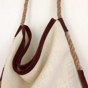 Peut inclure: Un sac cabas en toile blanche avec des anses en corde marron et une bordure en daim marron. Le sac est vide et suspendu &agrave; un mur blanc.