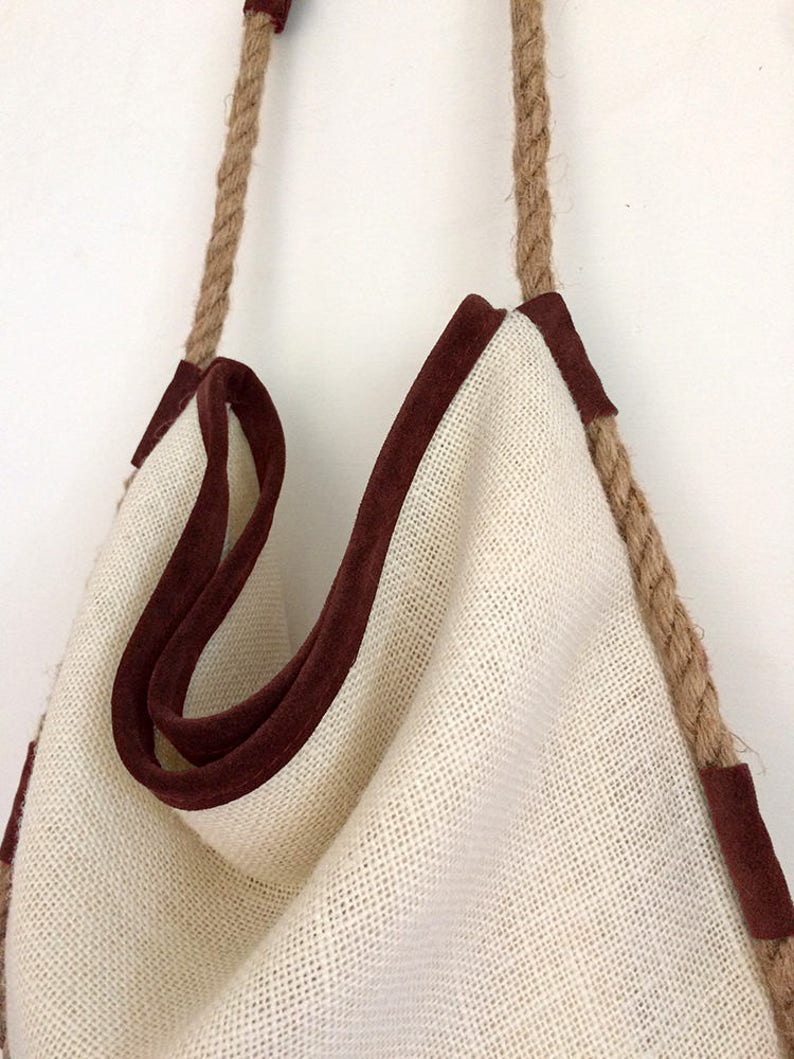 Peut inclure: Un sac cabas en toile blanche avec des anses en corde marron et une bordure en daim marron. Le sac est vide et suspendu &agrave; un mur blanc.