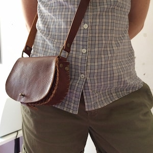 K&ouml;nnte beinhalten: Eine braune Leder-Umh&auml;ngetasche mit Klappenverschluss und einem geflochtenen Detail. Die Tasche wird &uuml;ber einem karierten Hemd und einer Khaki-Shorts getragen.