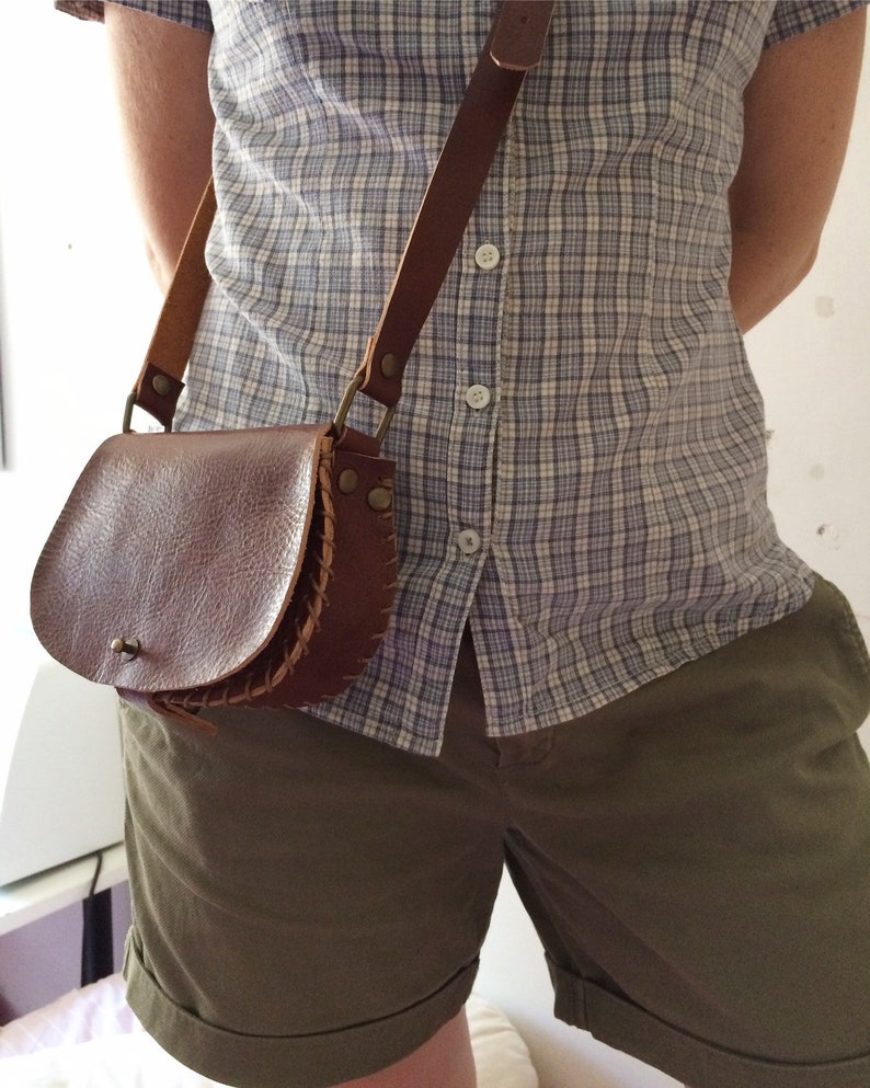 K&ouml;nnte beinhalten: Eine braune Leder-Umh&auml;ngetasche mit Klappenverschluss und einem geflochtenen Detail. Die Tasche wird &uuml;ber einem karierten Hemd und einer Khaki-Shorts getragen.