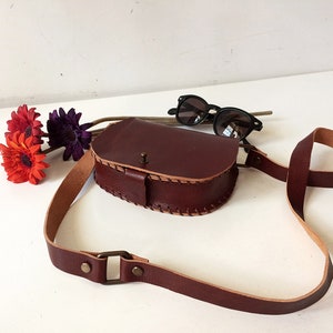 K&ouml;nnte beinhalten: Eine braune Leder-Umh&auml;ngetasche mit Klappenverschluss und einem langen, verstellbaren Trageriemen. Die Tasche hat ein gesticktes Design an den R&auml;ndern und eine kleine Metallschnalle am Riemen.