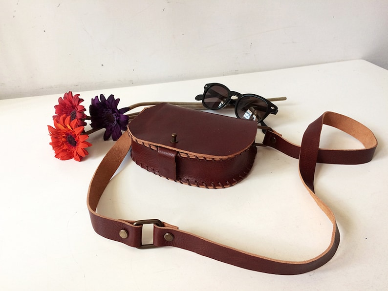 K&ouml;nnte beinhalten: Eine braune Leder-Umh&auml;ngetasche mit Klappenverschluss und einem langen, verstellbaren Trageriemen. Die Tasche hat ein gesticktes Design an den R&auml;ndern und eine kleine Metallschnalle am Riemen.
