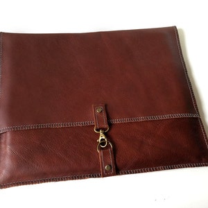 May include: A brown leather laptop sleeve with a flap closure and a brass clasp. The sleeve has a stitched edge and a subtle textured finish.