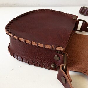K&ouml;nnte beinhalten: Eine braune Leder-Umh&auml;ngetasche mit Klappenverschluss und einem dekorativen Steppdetail. Die Tasche hat einen langen verstellbaren Riemen und eine Metallschnalle.