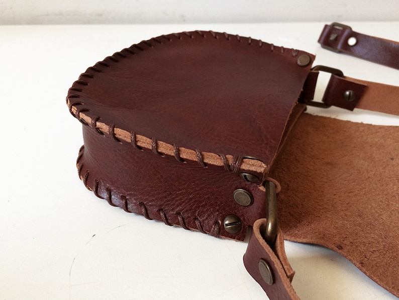 K&ouml;nnte beinhalten: Eine braune Leder-Umh&auml;ngetasche mit Klappenverschluss und einem dekorativen Steppdetail. Die Tasche hat einen langen verstellbaren Riemen und eine Metallschnalle.