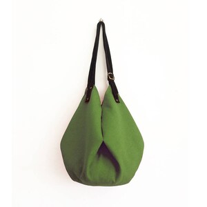 May include: A green canvas tote bag with black leather straps and a brass buckle. The bag is hanging from a hook on a white wall.