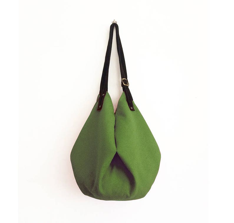 May include: A green canvas tote bag with black leather straps and a brass buckle. The bag is hanging from a hook on a white wall.
