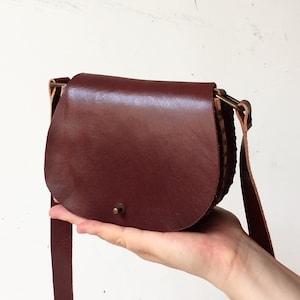 K&ouml;nnte beinhalten: Eine kleine braune Leder-Umh&auml;ngetasche mit Klappenverschluss und einer Messing-Schnalle. Die Tasche hat einen kurzen Riemen und einen langen verstellbaren Riemen.
