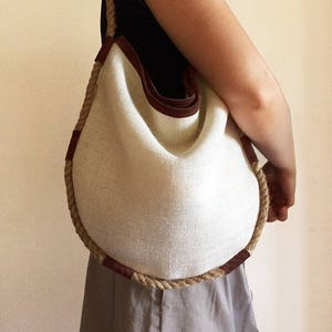 Peut inclure: Un sac cabas rond en tissu blanc tiss&eacute; avec des anses en corde marron et des accents en cuir marron. Le sac est tenu par une personne.