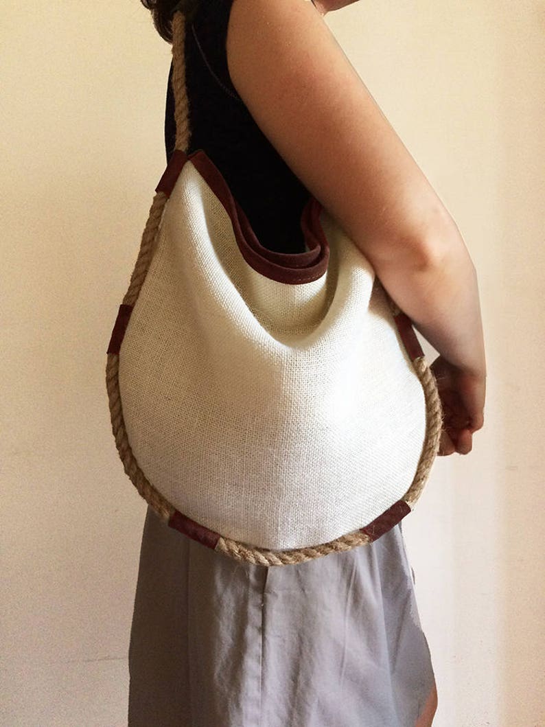 Peut inclure: Un sac cabas rond en tissu blanc tiss&eacute; avec des anses en corde marron et des accents en cuir marron. Le sac est tenu par une personne.