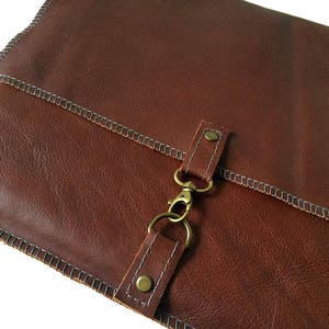 May include: A brown leather portfolio with a flap closure and a brass clasp. The portfolio has a stitched edge and a subtle textured surface.