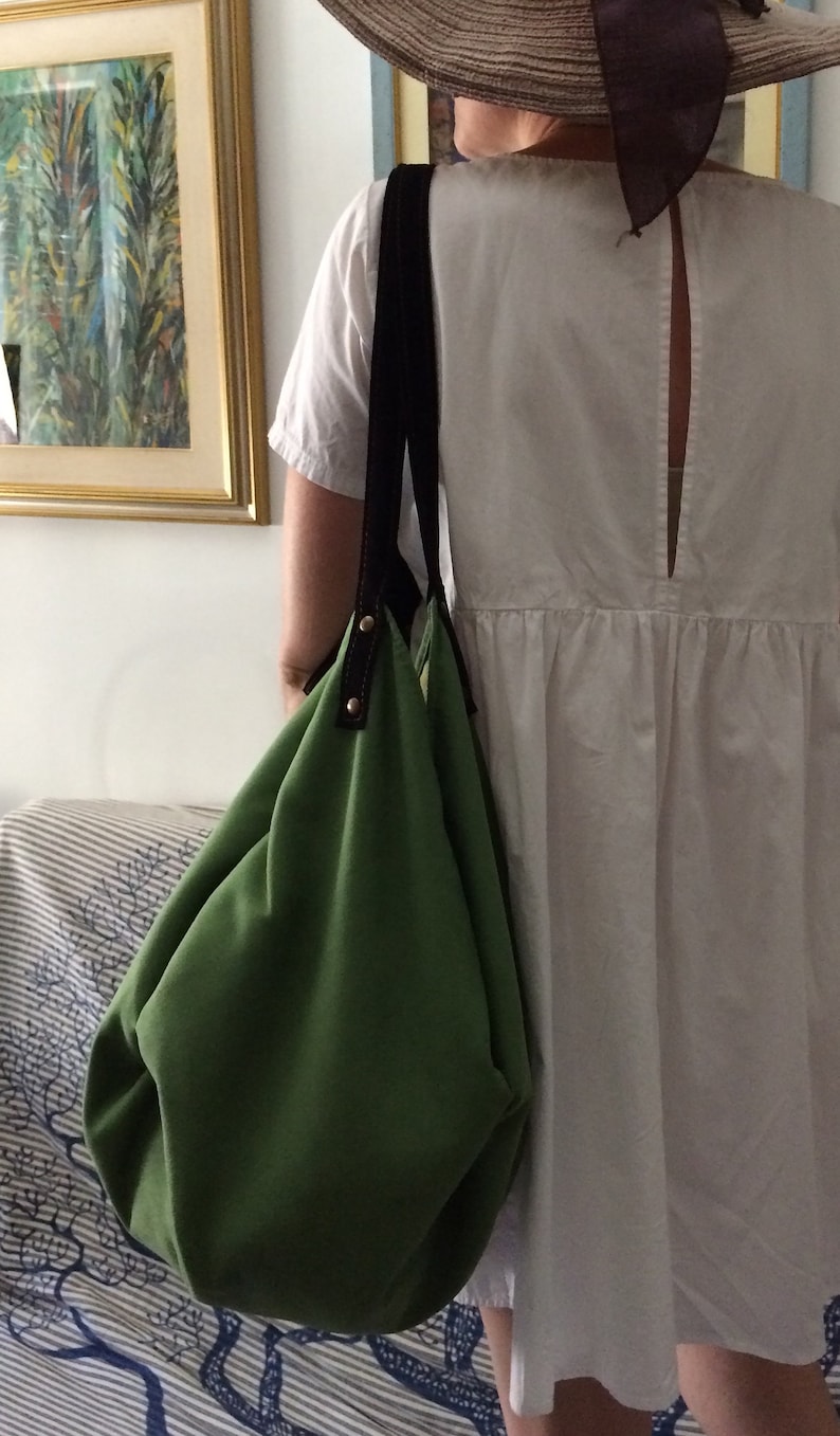May include: A green canvas tote bag with black straps and gold accents. The bag is being held by a person wearing a white dress and a straw hat.