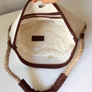 Peut inclure: Un sac cabas en toile blanche avec des anses en corde marron et une bordure en daim marron. Le sac a une poche int&eacute;rieure en tissu marron.