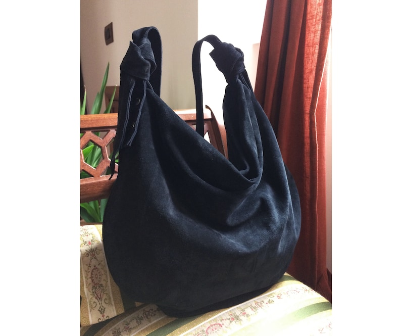 May include: A black suede hobo bag with a knotted handle and a long tassel. The bag is sitting on a patterned fabric surface.
