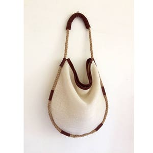 Peut inclure: Un sac cabas en toile blanche avec des anses en corde marron et des finitions en cuir marron. Le sac a une forme arrondie et est fait d'un tissu naturel.