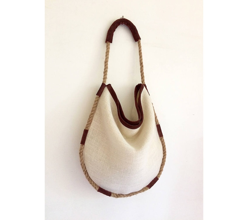 Peut inclure: Un sac cabas en toile blanche avec des anses en corde marron et des finitions en cuir marron. Le sac a une forme arrondie et est fait d'un tissu naturel.