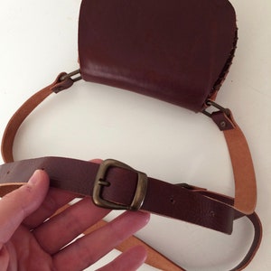K&ouml;nnte beinhalten: Eine braune Leder-Umh&auml;ngetasche mit einer Klappe und einer Messing-Schnalle. Die Tasche hat einen langen, verstellbaren Riemen.