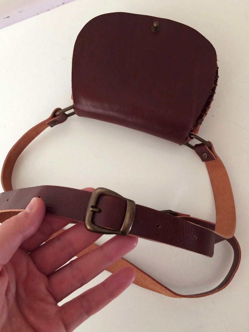K&ouml;nnte beinhalten: Eine braune Leder-Umh&auml;ngetasche mit einer Klappe und einer Messing-Schnalle. Die Tasche hat einen langen, verstellbaren Riemen.