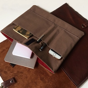 May include: A brown canvas bag with a leather flap and a red lining. The bag is open and has a phone, pens, and a notebook inside.