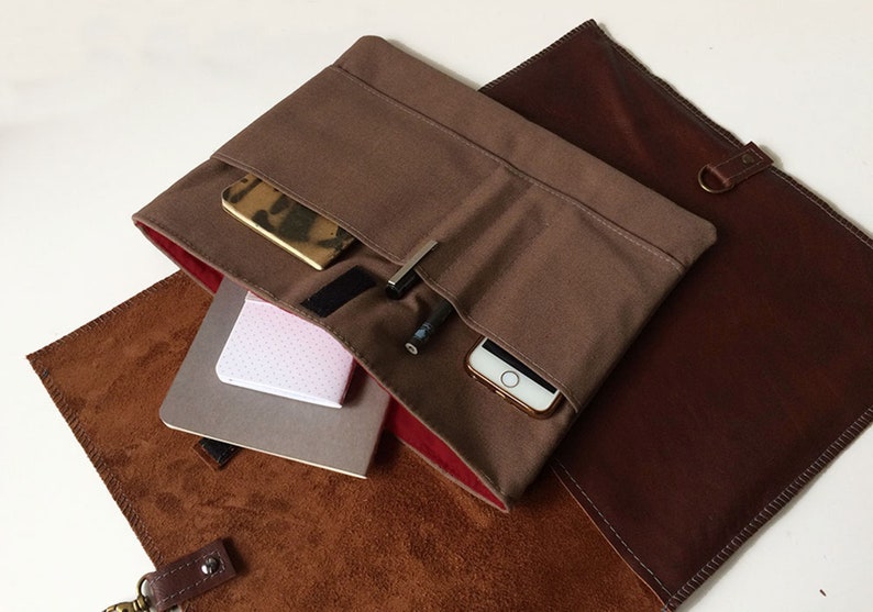 May include: A brown canvas bag with a leather flap and a red lining. The bag is open and has a phone, pens, and a notebook inside.