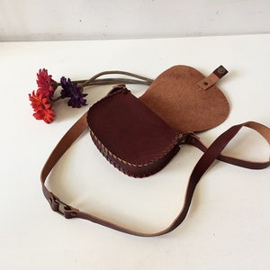 K&ouml;nnte beinhalten: Eine braune Leder-Umh&auml;ngetasche mit Klappenverschluss und einem langen, verstellbaren Trageriemen. Die Tasche ist offen und das Innere ist mit einem hellbraunen Stoff gef&uuml;ttert.