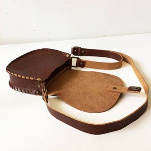 K&ouml;nnte beinhalten: Eine braune Leder-Umh&auml;ngetasche mit Klappenverschluss und einem langen, verstellbaren Trageriemen. Die Tasche hat ein gen&auml;htes Design und ein braunes Lederfutter.