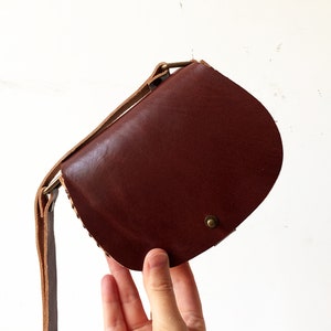 K&ouml;nnte beinhalten: Eine kleine braune Leder-Umh&auml;ngetasche mit einem beigefarbenen Riemen. Die Tasche hat eine abgerundete Form und einen Messingverschluss.