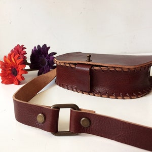 K&ouml;nnte beinhalten: Eine braune Leder-Umh&auml;ngetasche mit Klappenverschluss und einem gen&auml;hten Detail. Die Tasche hat einen langen verstellbaren Riemen und eine Messing-Schnalle.