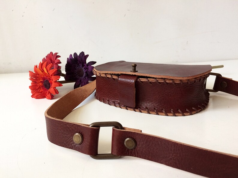 K&ouml;nnte beinhalten: Eine braune Leder-Umh&auml;ngetasche mit Klappenverschluss und einem gen&auml;hten Detail. Die Tasche hat einen langen verstellbaren Riemen und eine Messing-Schnalle.