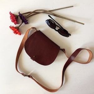 K&ouml;nnte beinhalten: Eine braune Leder-Umh&auml;ngetasche mit einem langen Riemen. Die Tasche hat eine abgerundete Form und einen kleinen Klappenverschluss. Die Tasche liegt auf einer wei&szlig;en Oberfl&auml;che mit Sonnenbrille und Blumen.