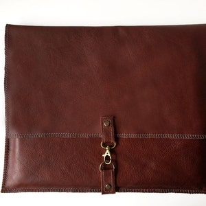 May include: A brown leather laptop sleeve with a flap closure and a metal clasp. The sleeve has a stitched design and a brown leather strap with a metal loop.
