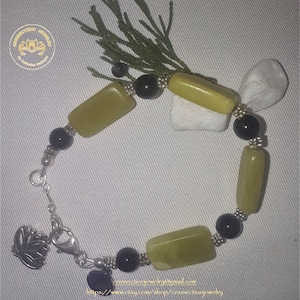 Puede incluir: Un brazalete verde y negro con un cierre plateado y un dije de loto. El brazalete está hecho con piedras verdes rectangulares y cuentas negras.