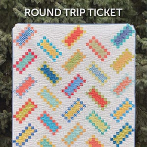 Könnte beinhalten: Ein farbenfrohes Patchwork-Quilt mit einem sich wiederholenden Muster aus rechteckigen Blöcken in verschiedenen Farben auf weißem Hintergrund. Der Quilt trägt den Text "ROUND TRIP TICKET" oben und "ATKINSON DESIGNS" unten. Ein Handarbeitsartikel.