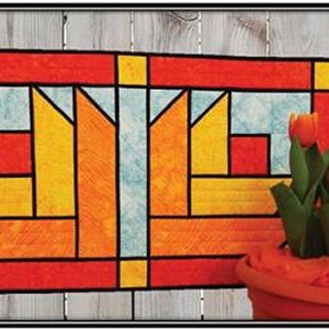 Könnte beinhalten: Ein farbenfrohes Quilt mit einem geometrischen Design in Rot-, Orange-, Gelb- und Blautönen. Das Quilt wird vor einem Holzgrund gezeigt, mit einem Topf mit orangefarbenen Tulpen im Vordergrund.
