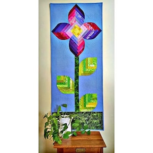May include: A vibrant, handmade quilt with a floral design. The flower has petals in shades of red, pink, purple, and blue, with a yellow centre. The stem and leaves are green. The quilt is displayed against a light blue backdrop.
