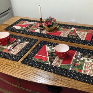 May include: Three red and green quilted placemats with black trim and gold accents, and a matching quilted table runner with a black border. The placemats and runner feature a patchwork design with various holiday-themed fabrics.