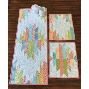 May include: A quilted table runner and two matching placemats with a colorful geometric design. The runner and placemats feature a white background with a pattern of pastel-colored fabric strips. A small white sugar bowl and creamer sit on the runner.
