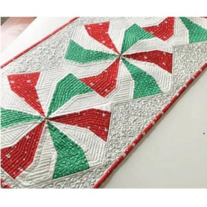 May include: A quilted table runner with a festive design. The runner features a repeating pattern of red, green, and white geometric shapes resembling pinwheels. The edges are trimmed with a red border, and the background is white.