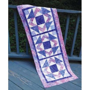 May include: A rectangular quilted table runner with a repeating pattern of blue and white geometric shapes on a pink and purple background. The runner is draped over a wooden railing.