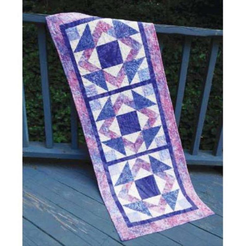 May include: A rectangular quilted table runner with a repeating pattern of blue and white geometric shapes on a pink and purple background. The runner is draped over a wooden railing.