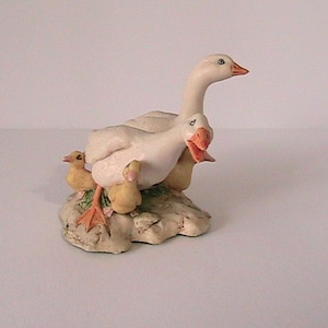 May include: A ceramic figurine of a goose family. The adult geese are white with orange beaks and feet, and the goslings are yellow. The geese are perched on a textured, light-colored base, creating a charming decorative piece.