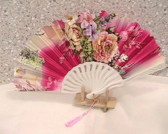 May include: A decorative hand fan with a white plastic handle and a pink tassel. The fan's fabric is adorned with a floral design featuring roses and other flowers in shades of pink, purple, and cream.