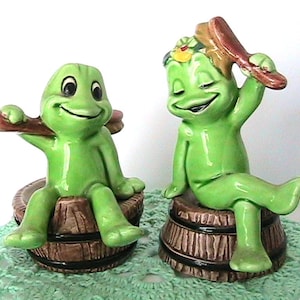 May include: Two ceramic frog figurines. Each green frog is sitting on a brown barrel. One frog holds a brown object over its shoulder, and the other holds a brown object over its head. The frogs have smiling faces.