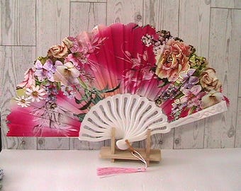 May include: A decorative hand fan with a white handle and a vibrant floral design. The fan features a pink and red background with detailed illustrations of various flowers in shades of pink, purple, and cream.