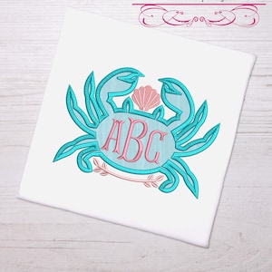 May include: A turquoise blue crab with a pink seashell on top and a circle with the letters ABC inside. The crab is surrounded by a leafy design.