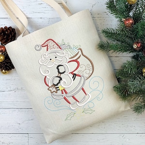 May include: Beige canvas tote bag featuring an embroidered Santa Claus design. The Santa Claus illustration includes a red hat, white beard, and a bag of gifts. The bag has beige handles and is suitable for carrying items.